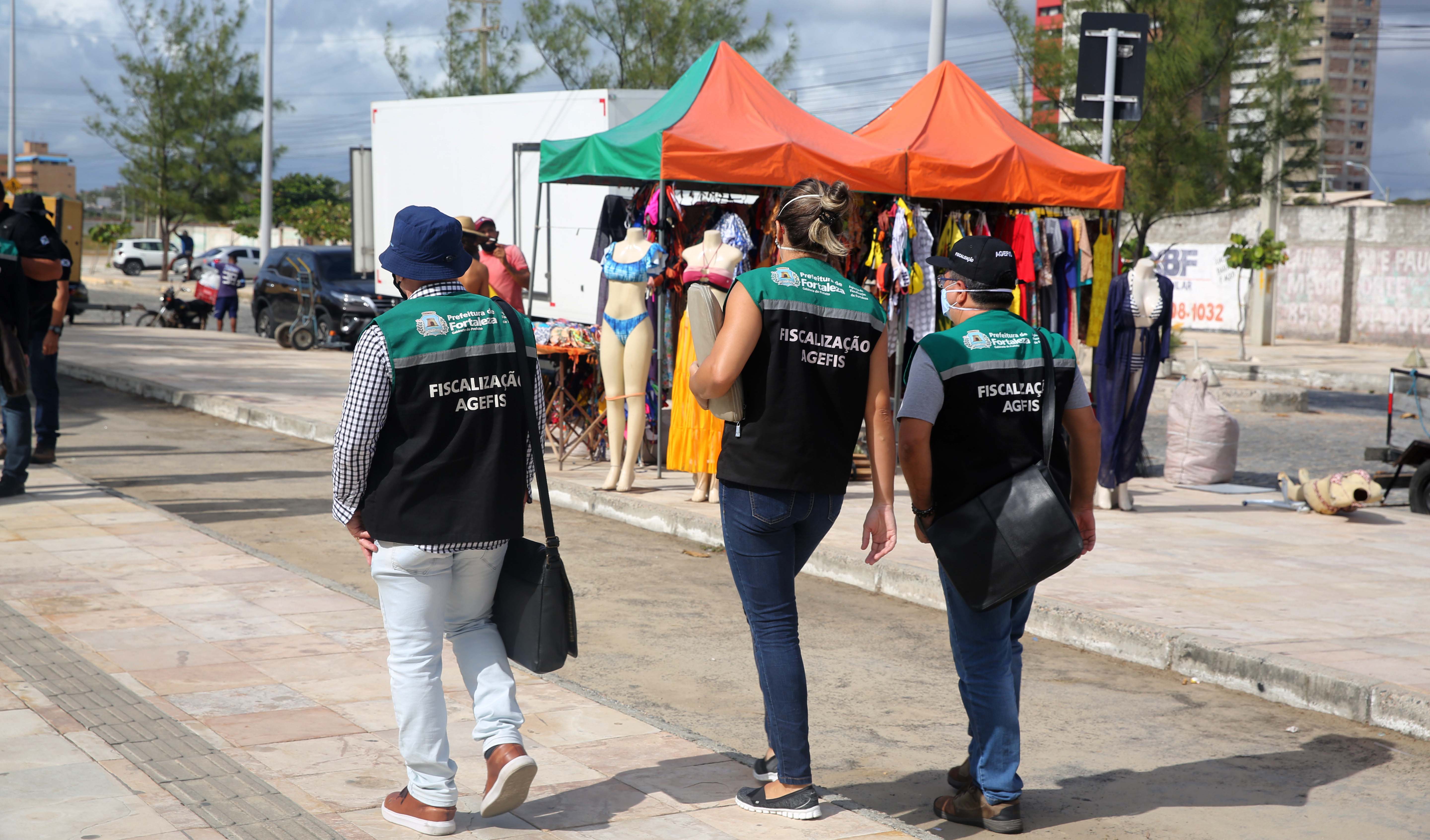 a foto mostra fiscais da agefis, que são identificados pelos coletes verdes andando pelo calçadão da praia do futuro, eles estão de costas